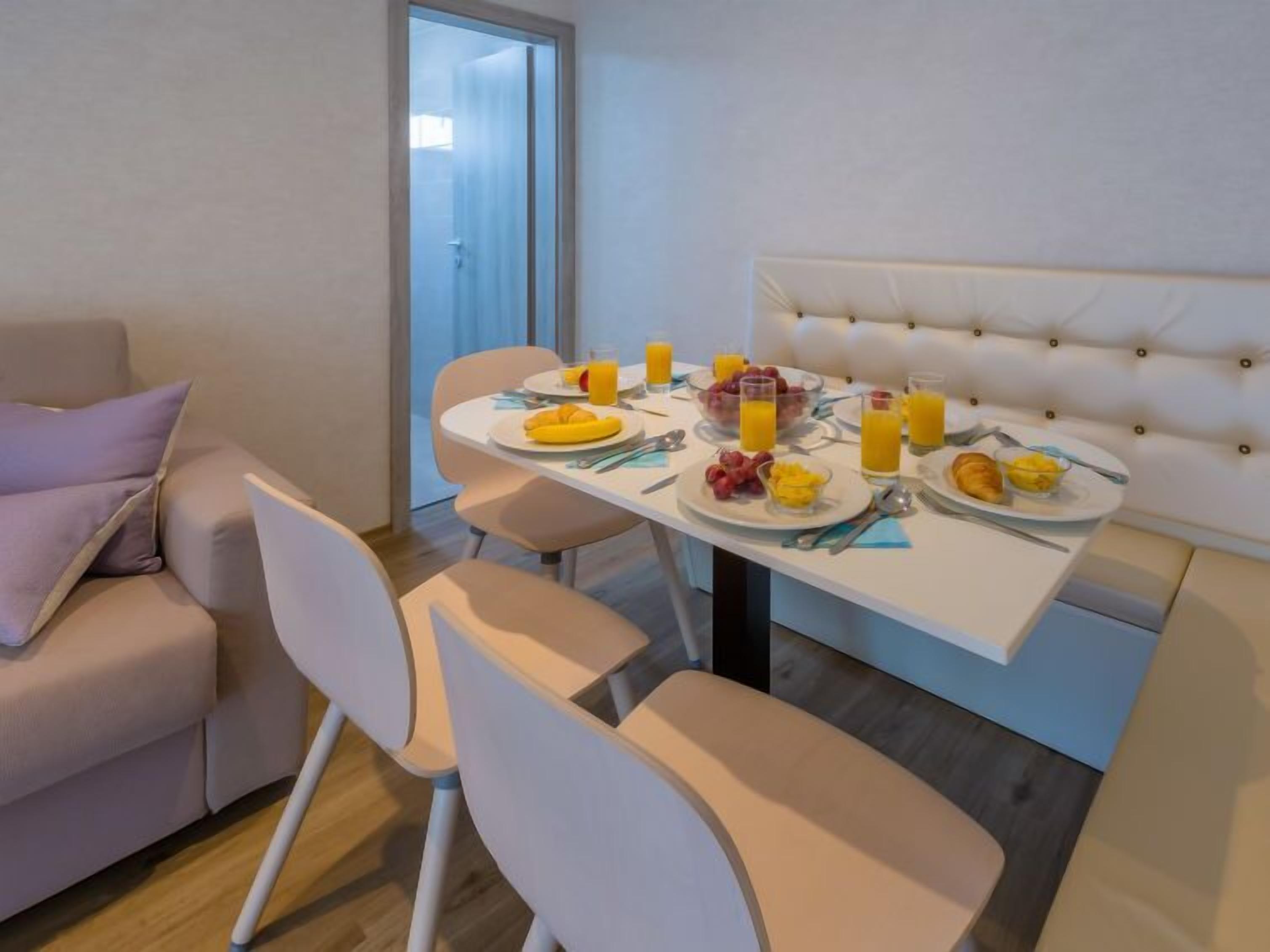 A dining area with a white table set for breakfast, including plates of fruit and juice, and a sofa with cushions.