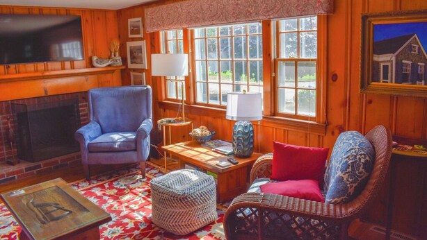 A cozy living room with a fireplace, comfortable seating, and large windows offering a view of the outdoors. - Lovely 3 bedroom ranch home well maintained by owners (Dennis Port)