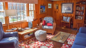 A cozy room with wooden walls, a blue armchair with red cushions, a wicker sofa, a small round ottoman, and a bookshelf filled with books. - Lovely 3 bedroom ranch home well maintained by owners (Dennis Port)