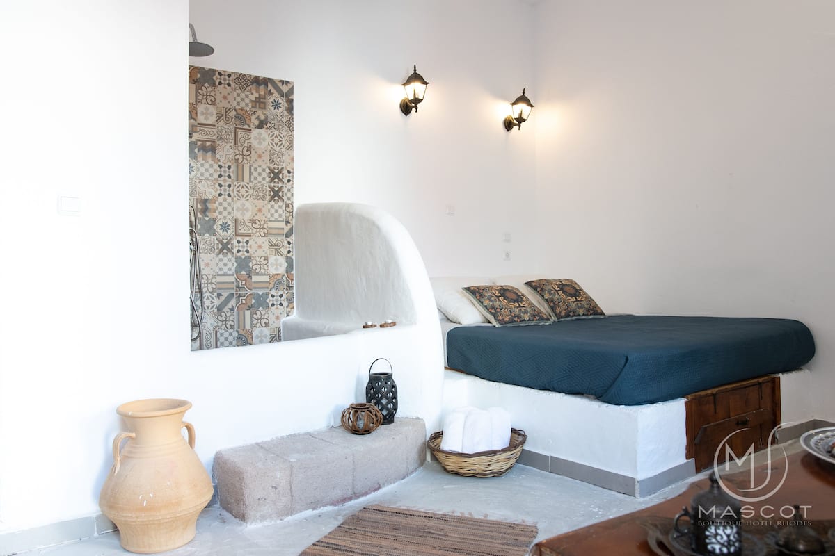 A bedroom with a bed, a wall-mounted light fixture, a decorative wall piece, a vase, and a basket with towels.