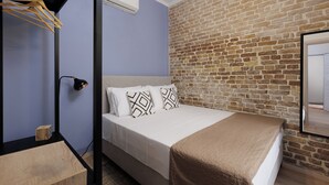 Une chambre avec un mur en briques, un lit agrémenté d’oreillers à motifs, une table de chevet en bois, une lampe de bureau noire et un miroir sur le mur.