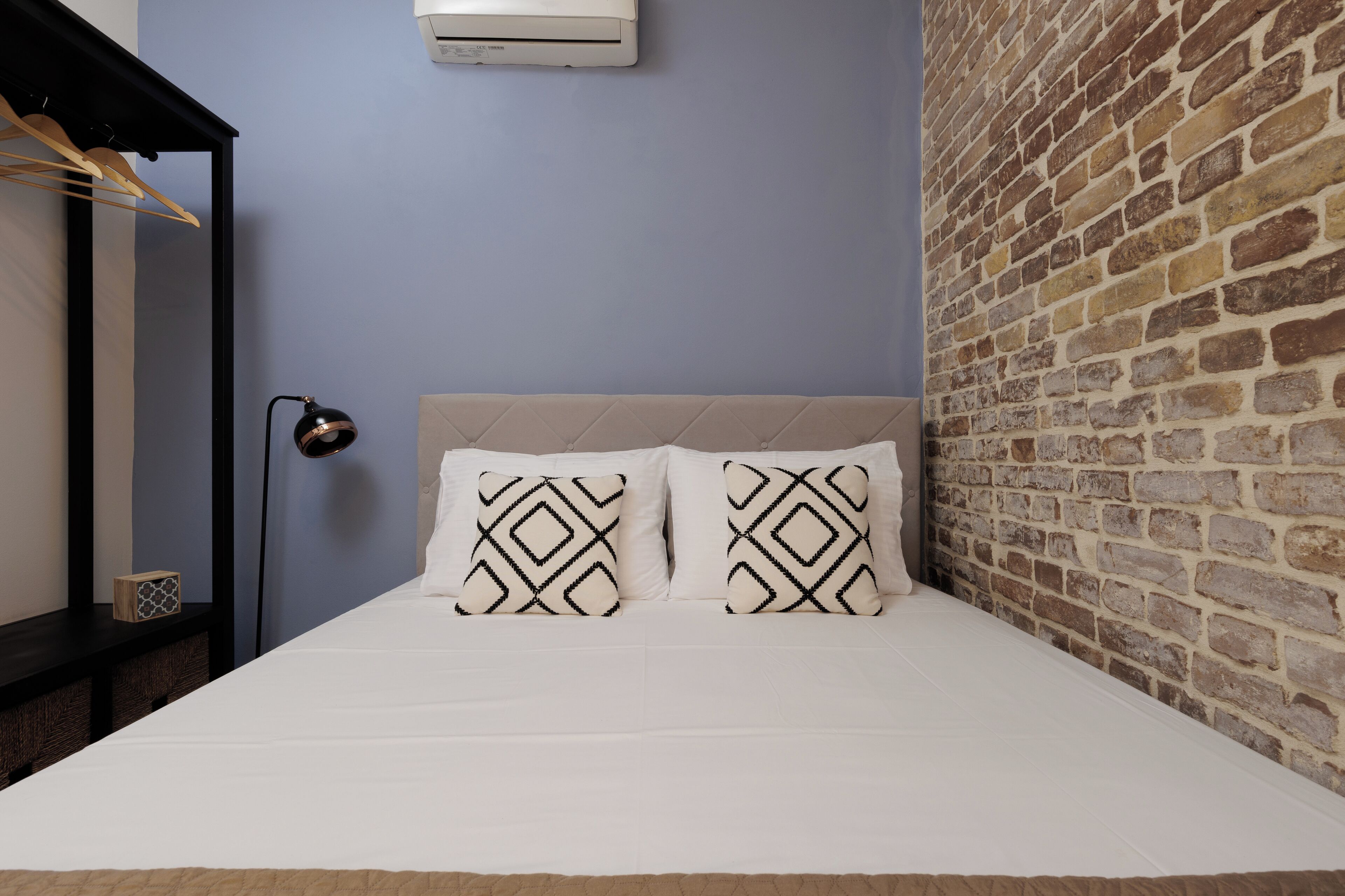 A bed with a beige headboard, two patterned pillows, a bedside lamp, and a brick wall.