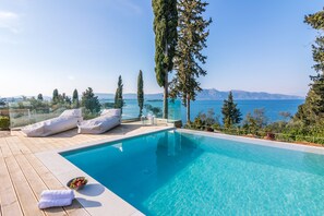 A pool with clear blue water, surrounded by a deck with white cushions and a bowl of fruit. - Melithea Seaview Villa (Corfu)