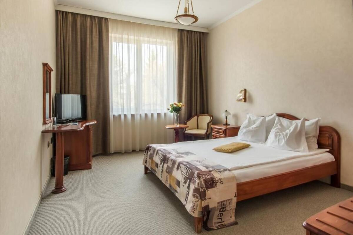 A hotel room with a bed, a desk with a television, a chair, a nightstand, and a window with curtains.