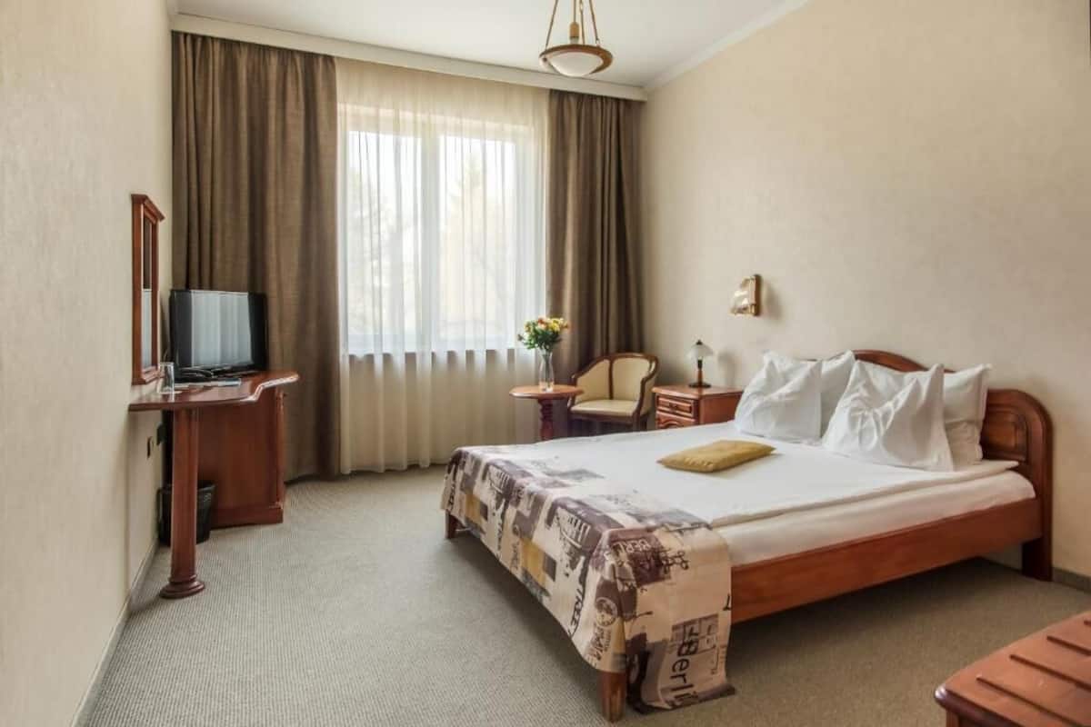 A hotel room with a bed, a desk with a television, a chair, a nightstand, and a window with curtains.