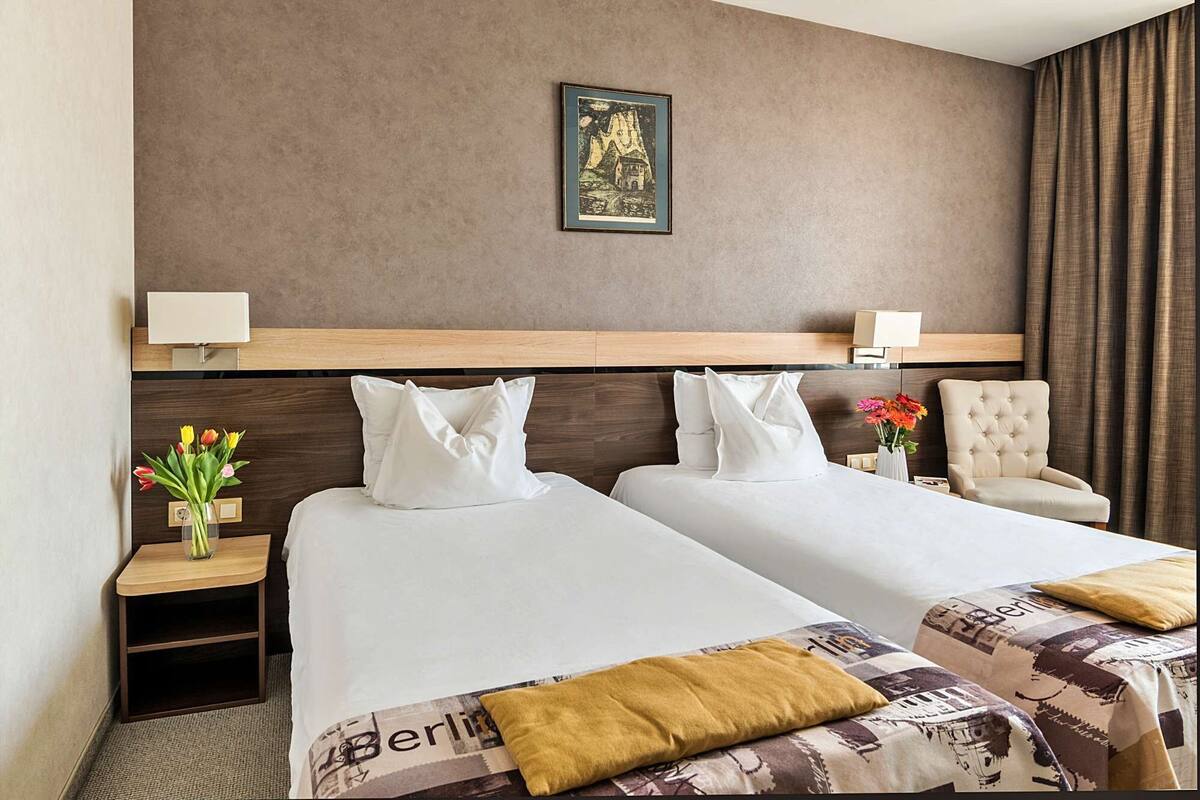 A hotel room with two beds, a wooden headboard, a framed picture on the wall, a nightstand with a lamp, a chair, and a vase with flowers.
