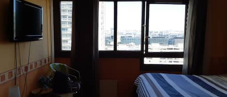 A hotel room with a bed, a television mounted on the wall, a desk with a lamp, and a window offering a city view.