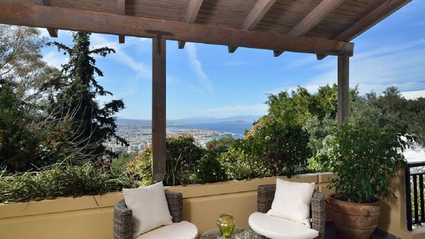 A covered outdoor seating area with wicker chairs and a glass table, overlooking a cityscape and greenery.