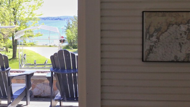 A view from a room with a window overlooking a grassy area and a body of water. There are two Adirondack chairs and a table outside.