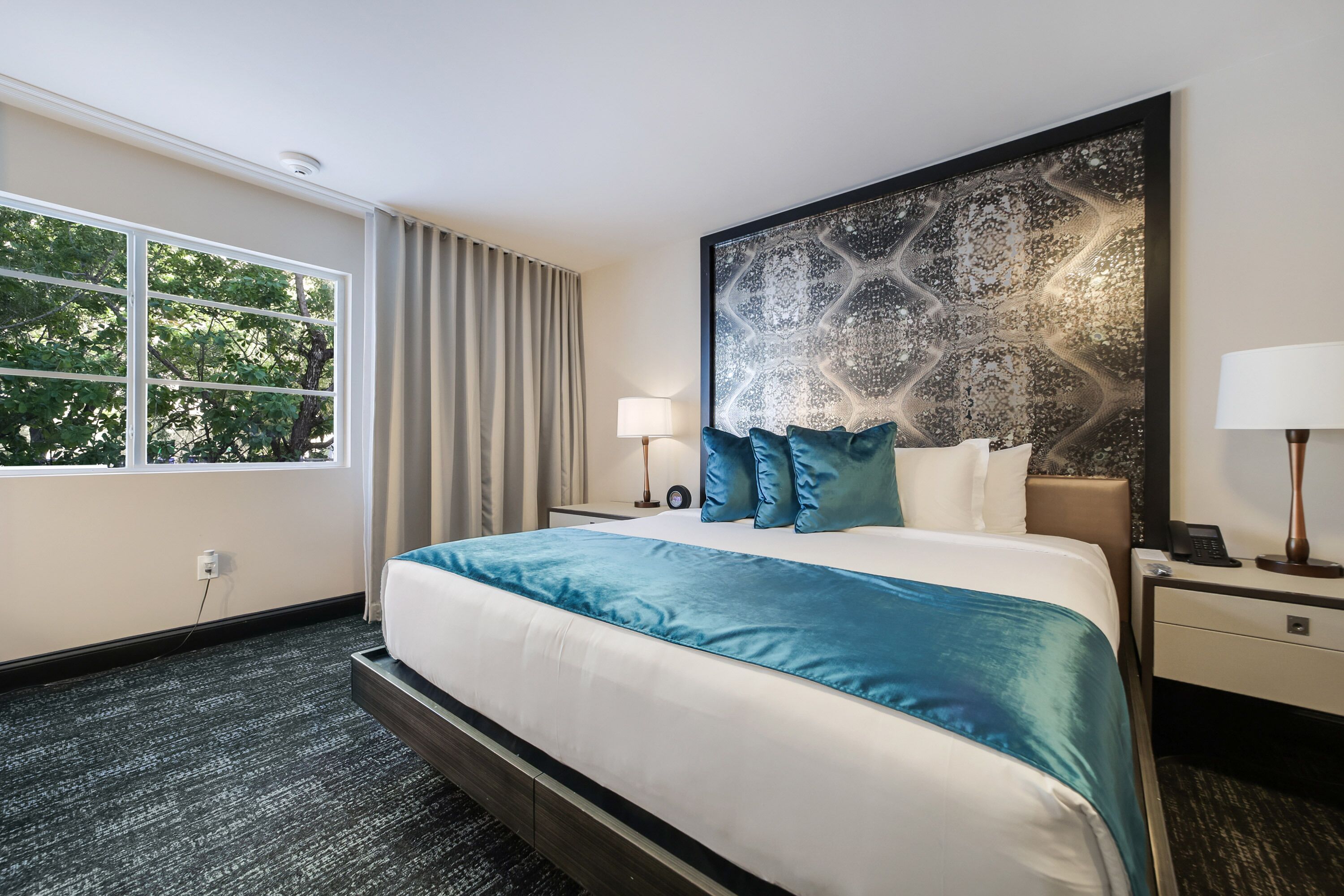 Una habitación de hotel con una cama grande, un cabecero con un panel decorativo, una mesita de noche con una lámpara y una ventana con cortinas.
