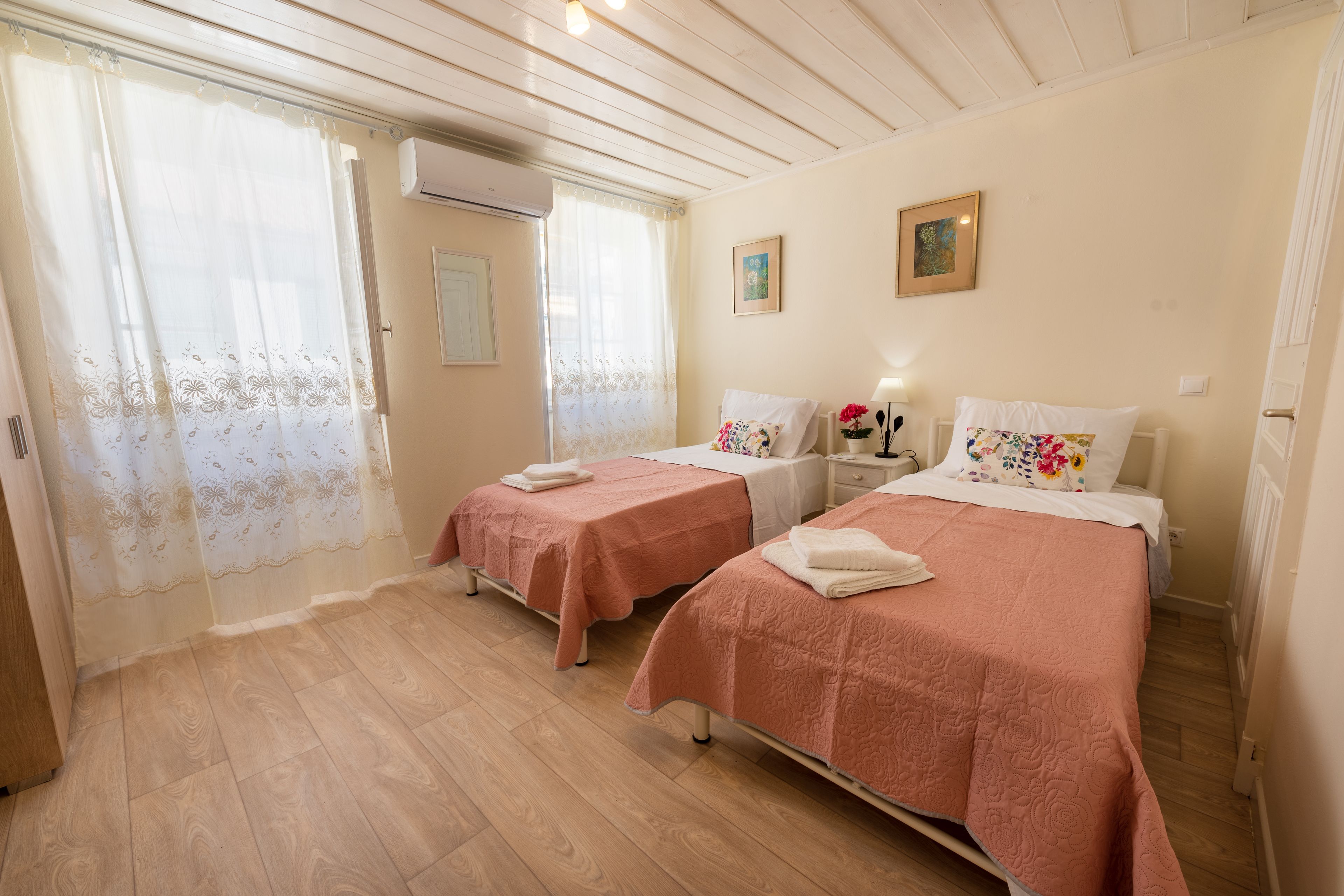 Une chambre avec deux lits, chacun recouvert d’une couvre-lit rose et agrémenté d’oreillers blancs, une petite table de chevet avec une lampe, et une fenêtre ornée de rideaux légers.
