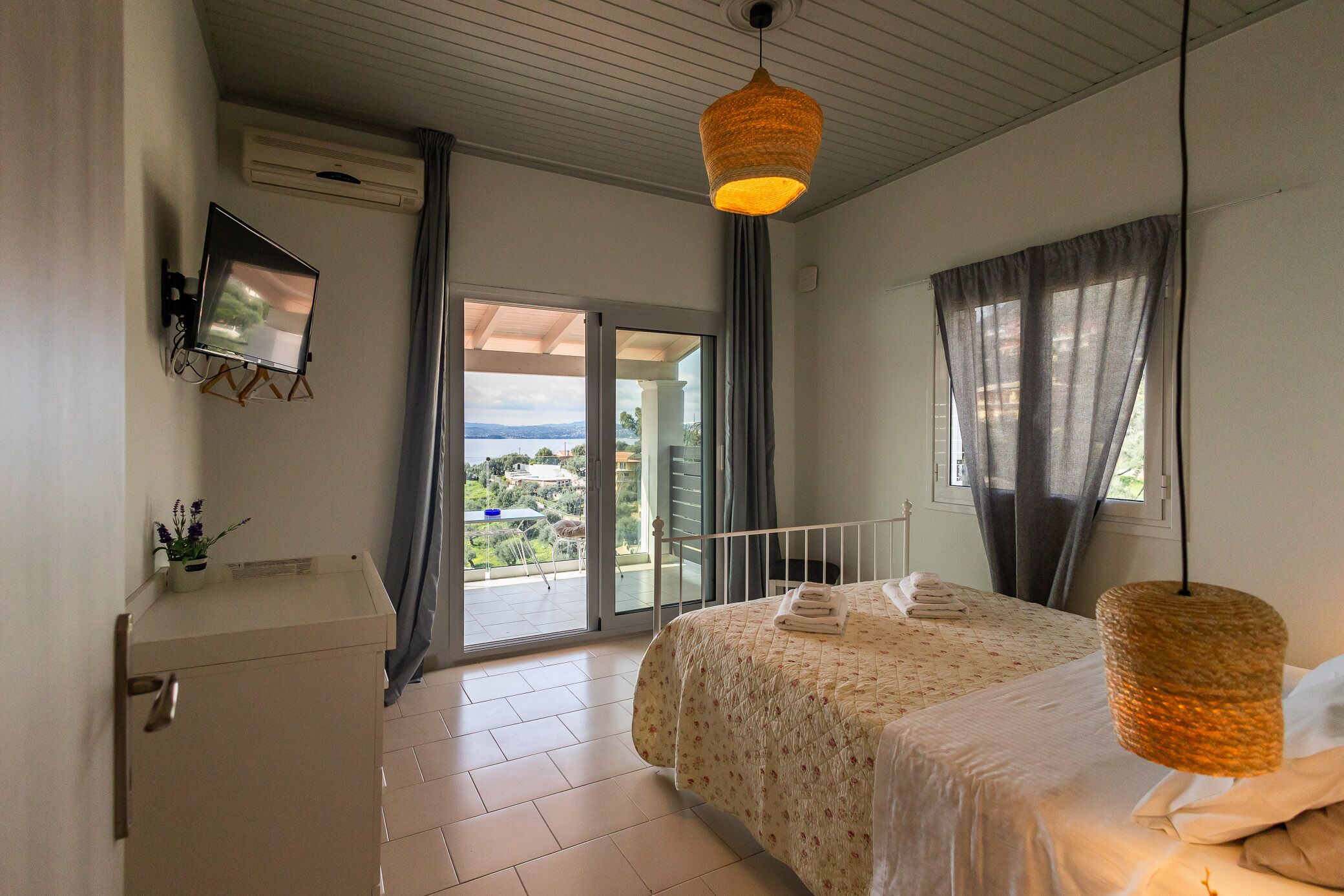 A bedroom with a bed, a bedside table, a TV mounted on the wall, and a balcony with a view.