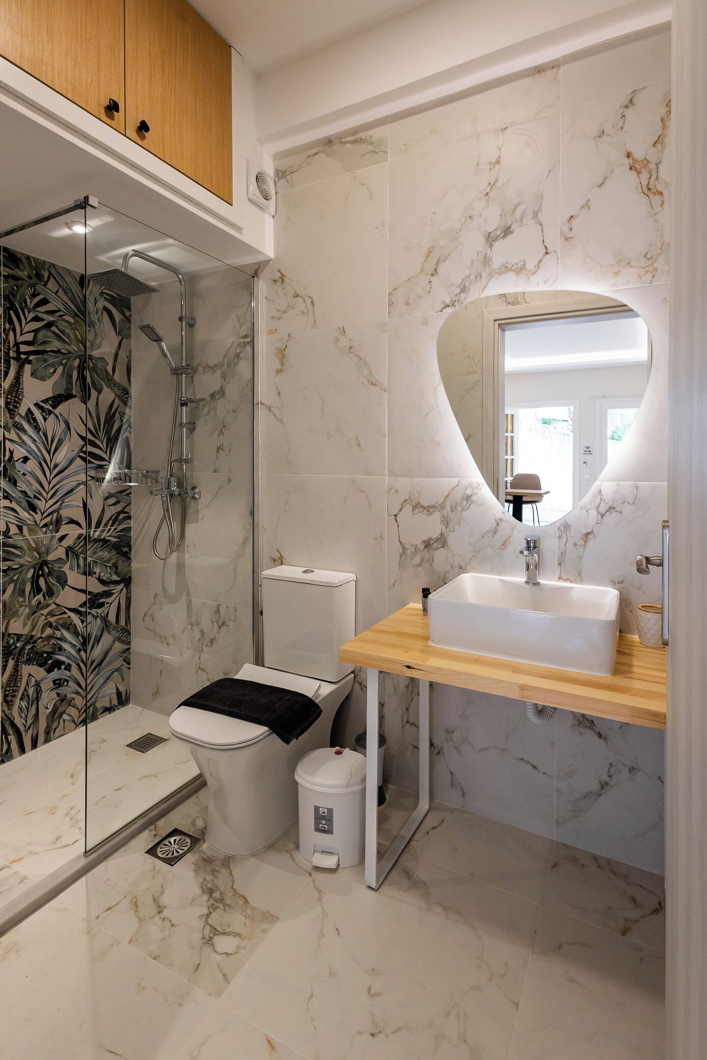 Une salle de bain dotée d’une douche à parois de verre, de toilettes blanches, d’un lavabo posé sur un plan de travail en bois et d’un mur recouvert de carreaux de marbre.