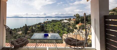 A balcony with a glass table and two cushioned chairs, overlooking a lake and hilly landscape.