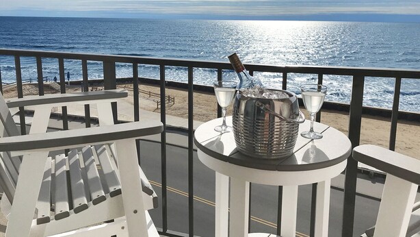 A balcony with a table set for two, overlooking a beach with the ocean in the background. - Beachfront 1Br/1Ba condo, free parking & WiFi  w/ Heated Pool (North Wildwood)
