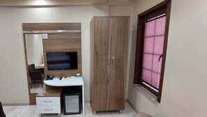 Una habitación compacta con un armario de madera, un escritorio blanco con cajones, un refrigerador pequeño, un televisor montado en la pared y una ventana con persianas.