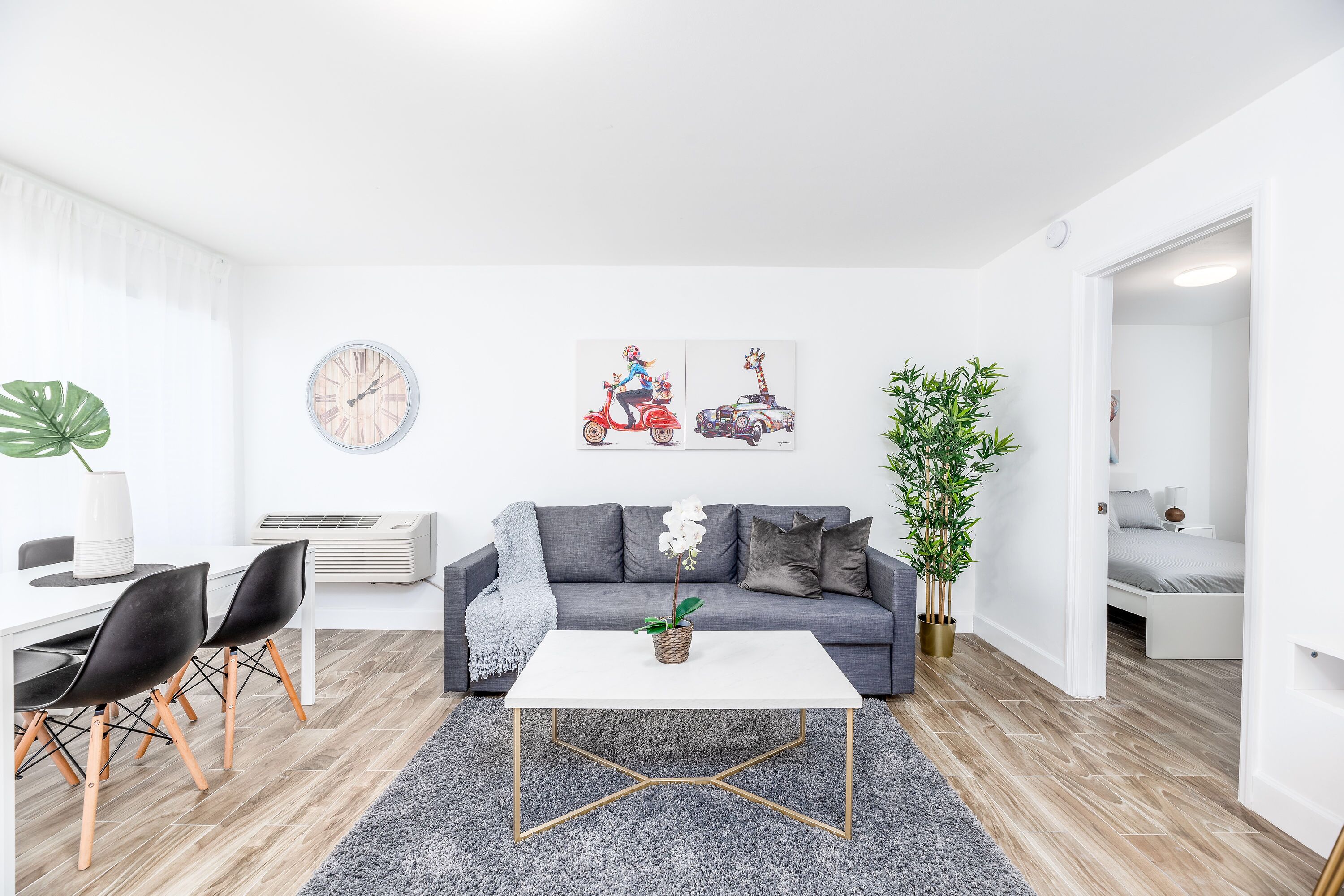 A modern living room with a grey sofa, a white coffee table, and a dining area with black chairs. There are two wall-mounted clocks and a potted plant.