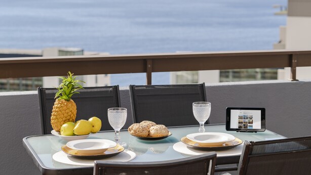 Une table dressée avec des assiettes, des verres, un ananas, des citrons et du pain, sur un balcon offrant une vue sur l’océan.