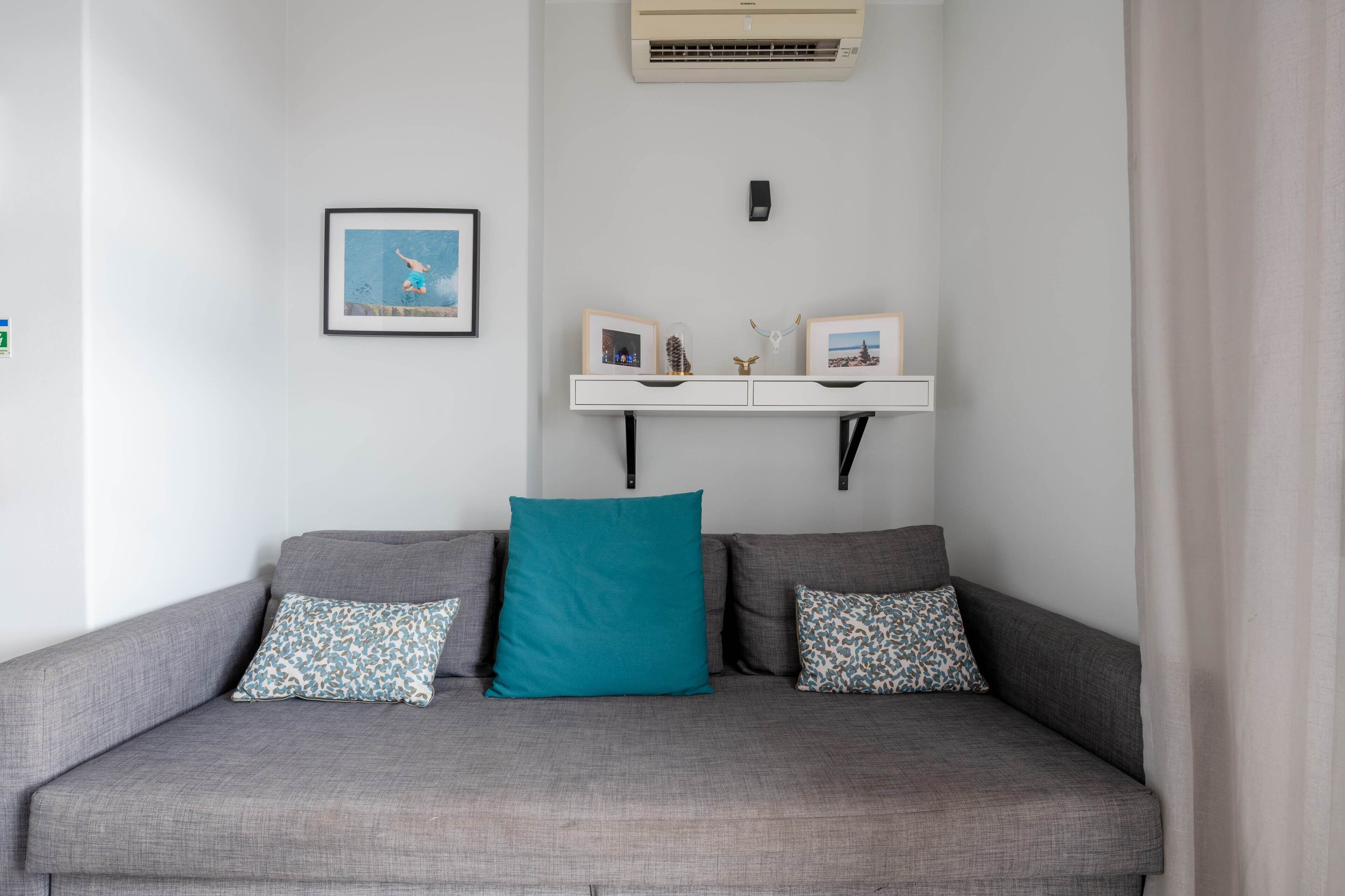 Un canapé gris agrémenté de coussins turquoise et à motifs, une étagère blanche ornée de photos encadrées et d’objets décoratifs, et un climatiseur fixé au mur.