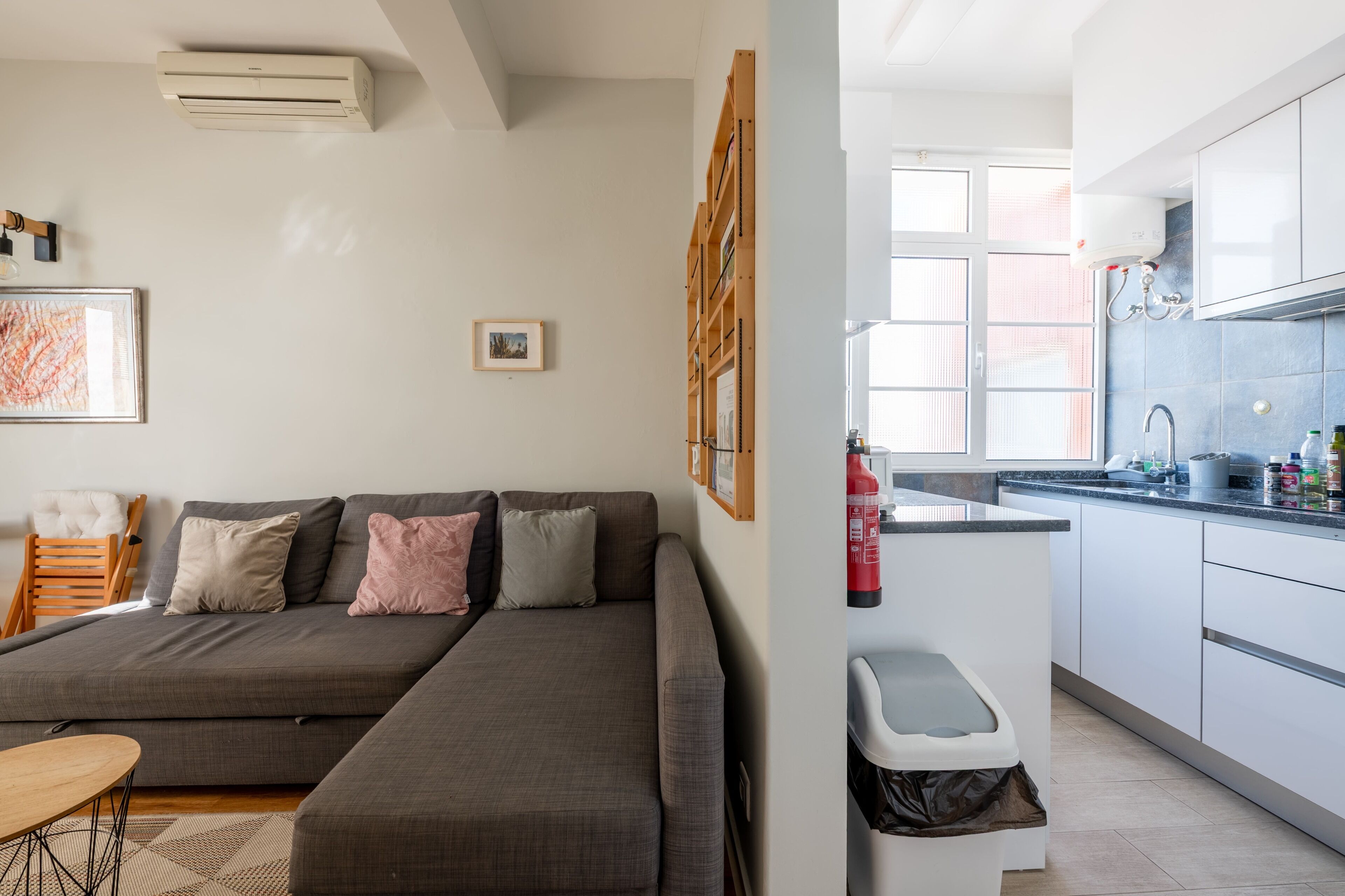 Una sala de estar moderna con un sofá gris, una mesita de madera y una cocina con armarios blancos y encimera negra.