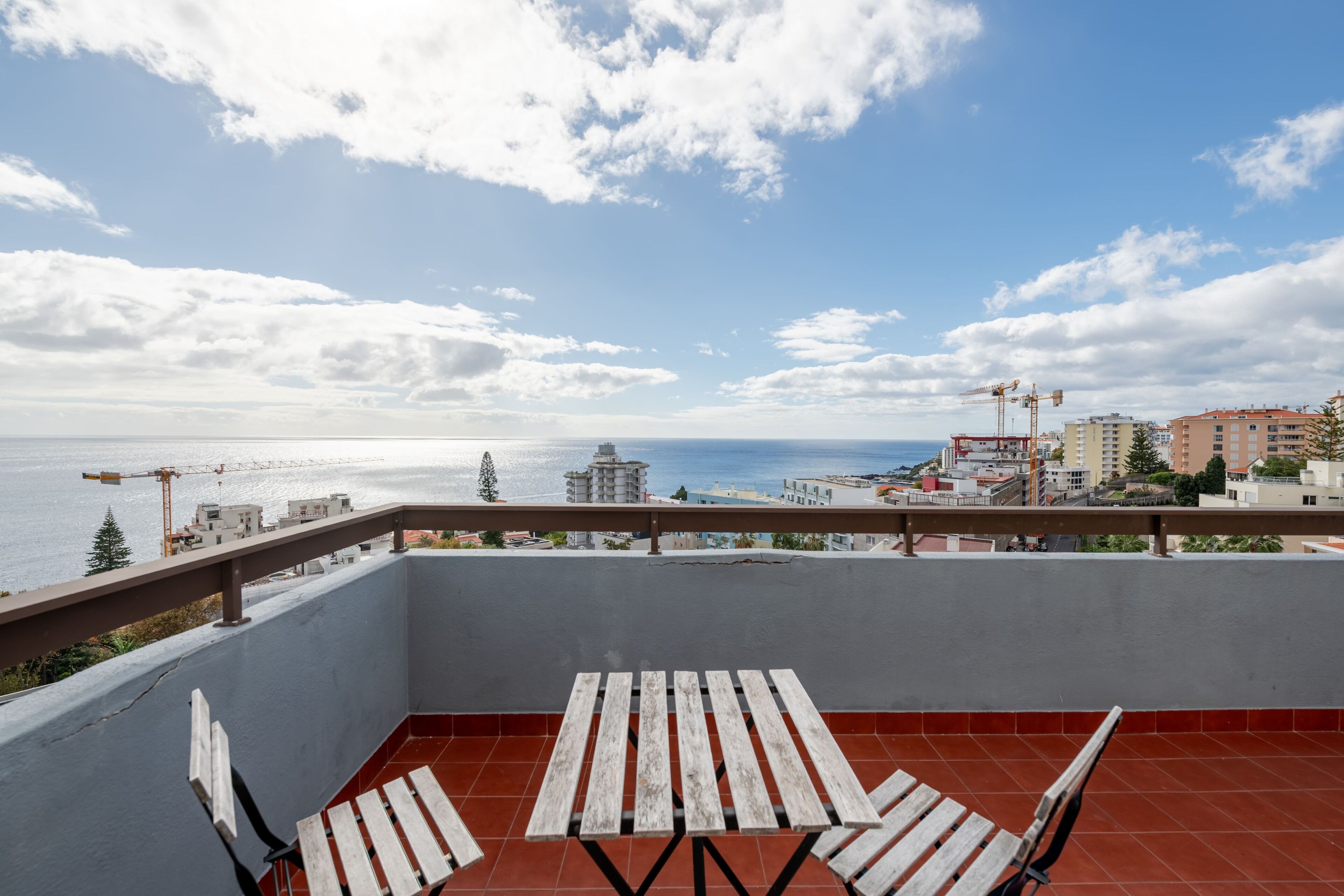 Een balkon met een houten tafel en stoelen, met uitzicht op een kustplaats en een heldere lucht.