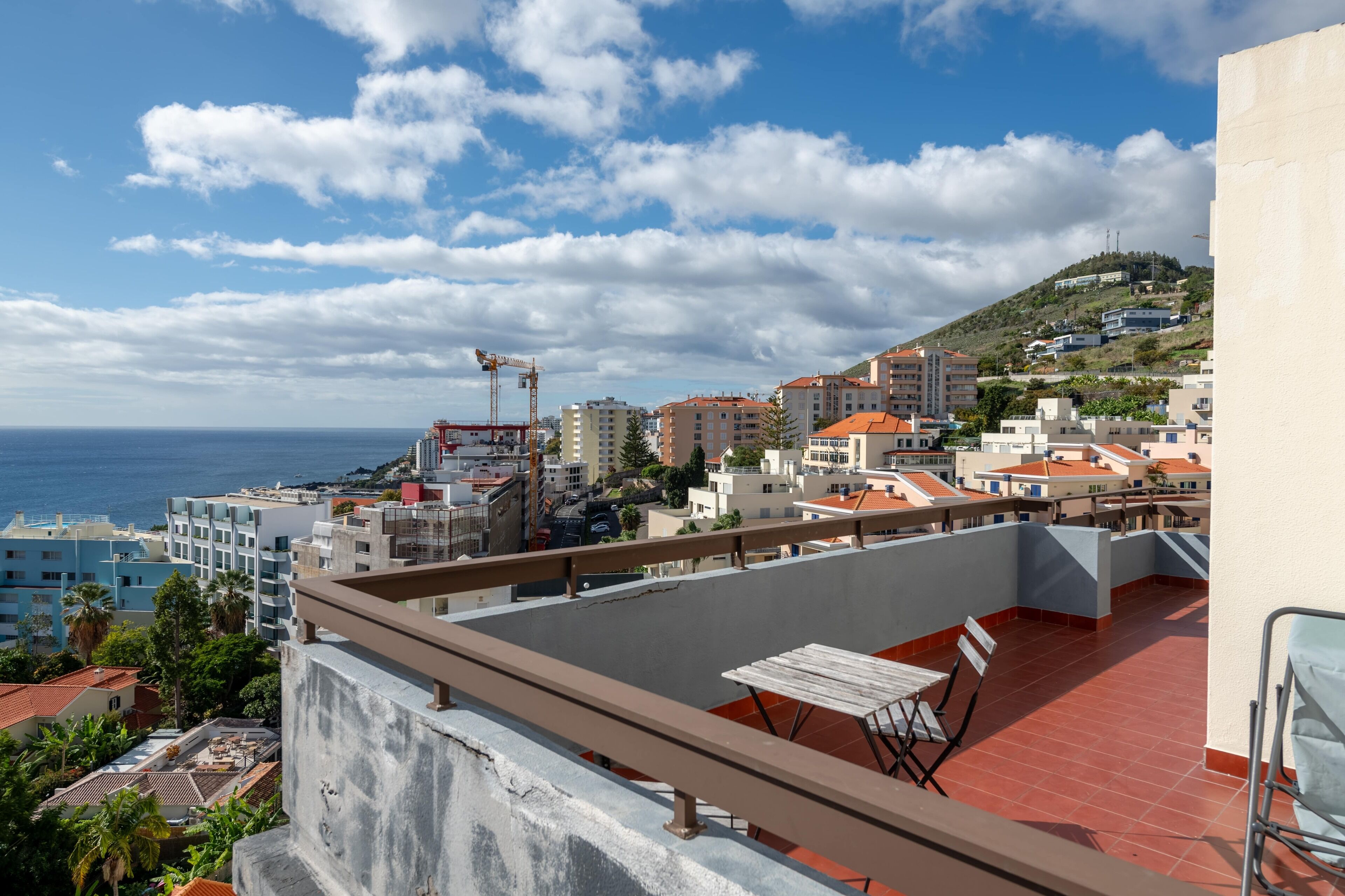 A rooftop terrace with a table and chairs, overlooking a coastal cityscape with buildings and a construction crane.