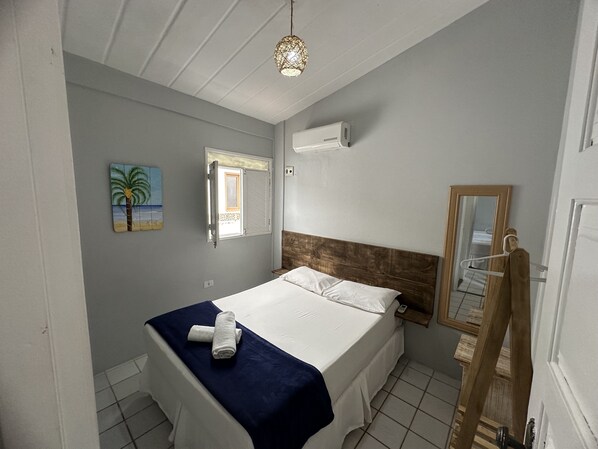 Une chambre à coucher comprenant un lit, une fenêtre, un climatiseur fixé au mur, une serviette, un tableau et un cintre en bois.