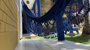 A hammock hanging between a yellow brick wall and a blue structure, with a tiled floor and a sign in the background. - Pousada Girassol (Ipojuca)
