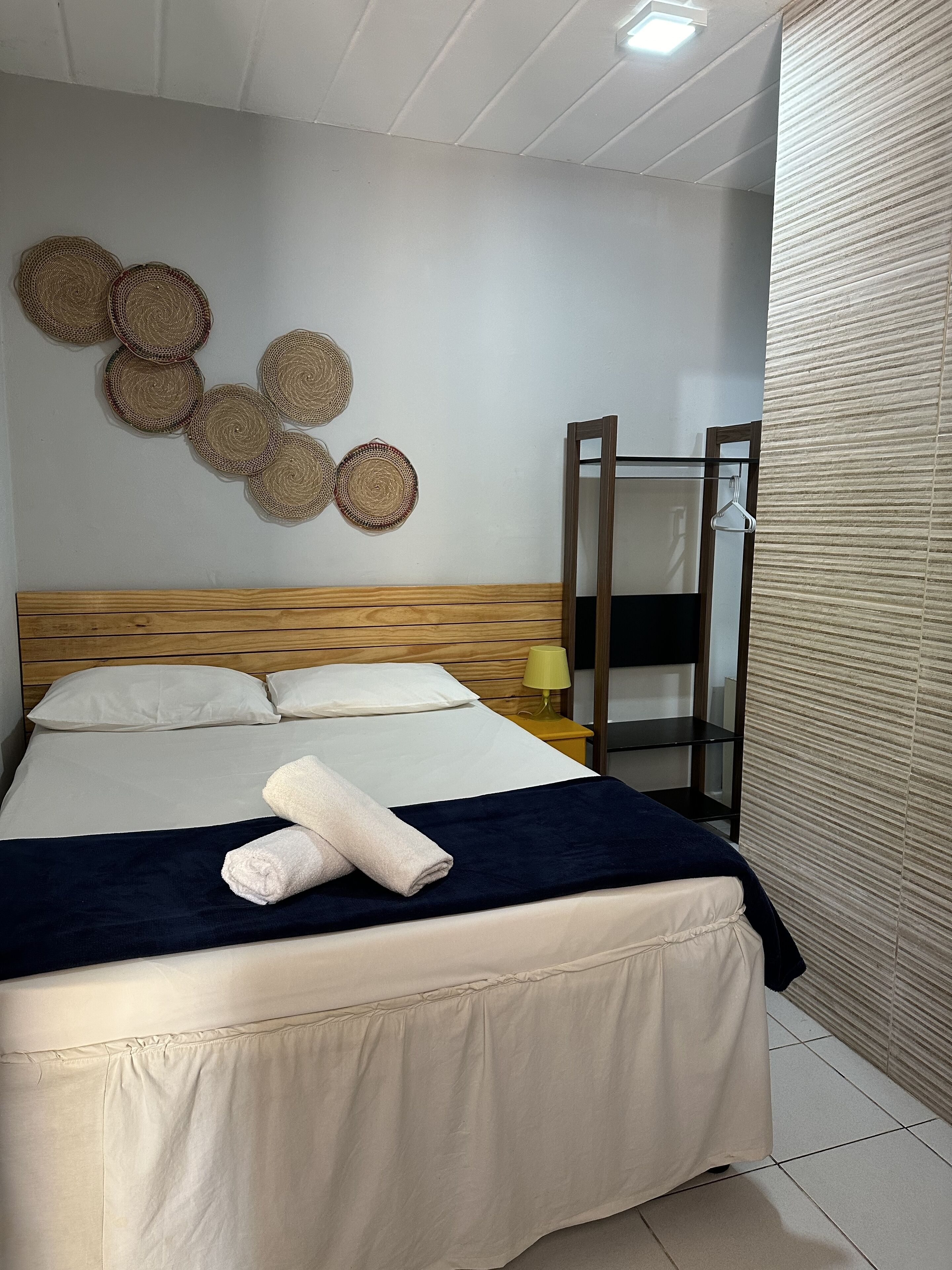 Un lit bien fait, avec une tête de lit en bois, une couvre-lit bleu foncé et deux serviettes enroulées. On peut voir des décorations murales tissées et un miroir avec un crochet fixé au mur.