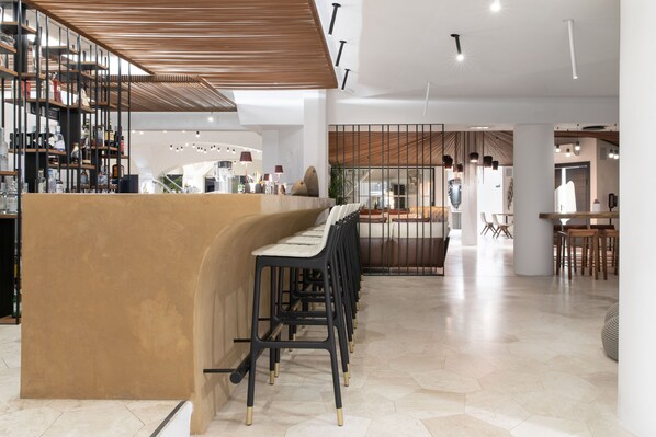 Uma área de bar moderna com balcão curvo e vários bancos. Há uma escada que leva a um andar superior, e uma área de estar com mesas e cadeiras ao fundo.