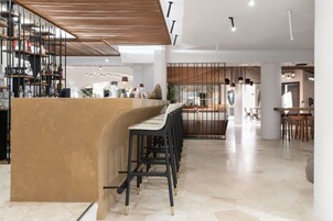 Un bar moderno con una barra curva y varios taburetes. Se ve una escalera que sube a un nivel superior, y un área de estar con mesas y sillas al fondo.