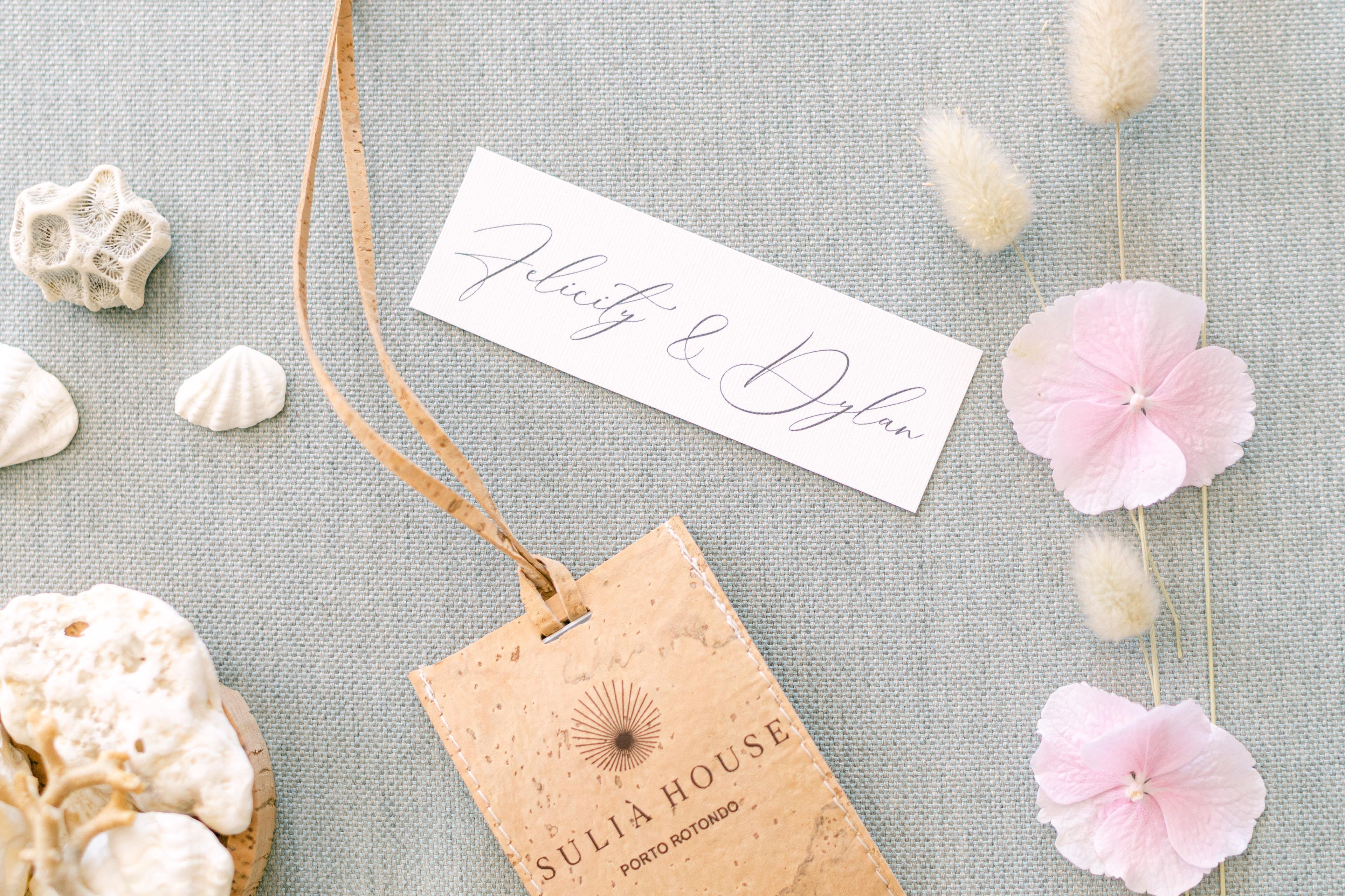Una tarjeta de identificación con la leyenda 'Jelicity & Dylan', una etiqueta que dice 'SULIA HOUSE PORTO ROTONDO', y caracolas y flores decorativas sobre un fondo texturizado.