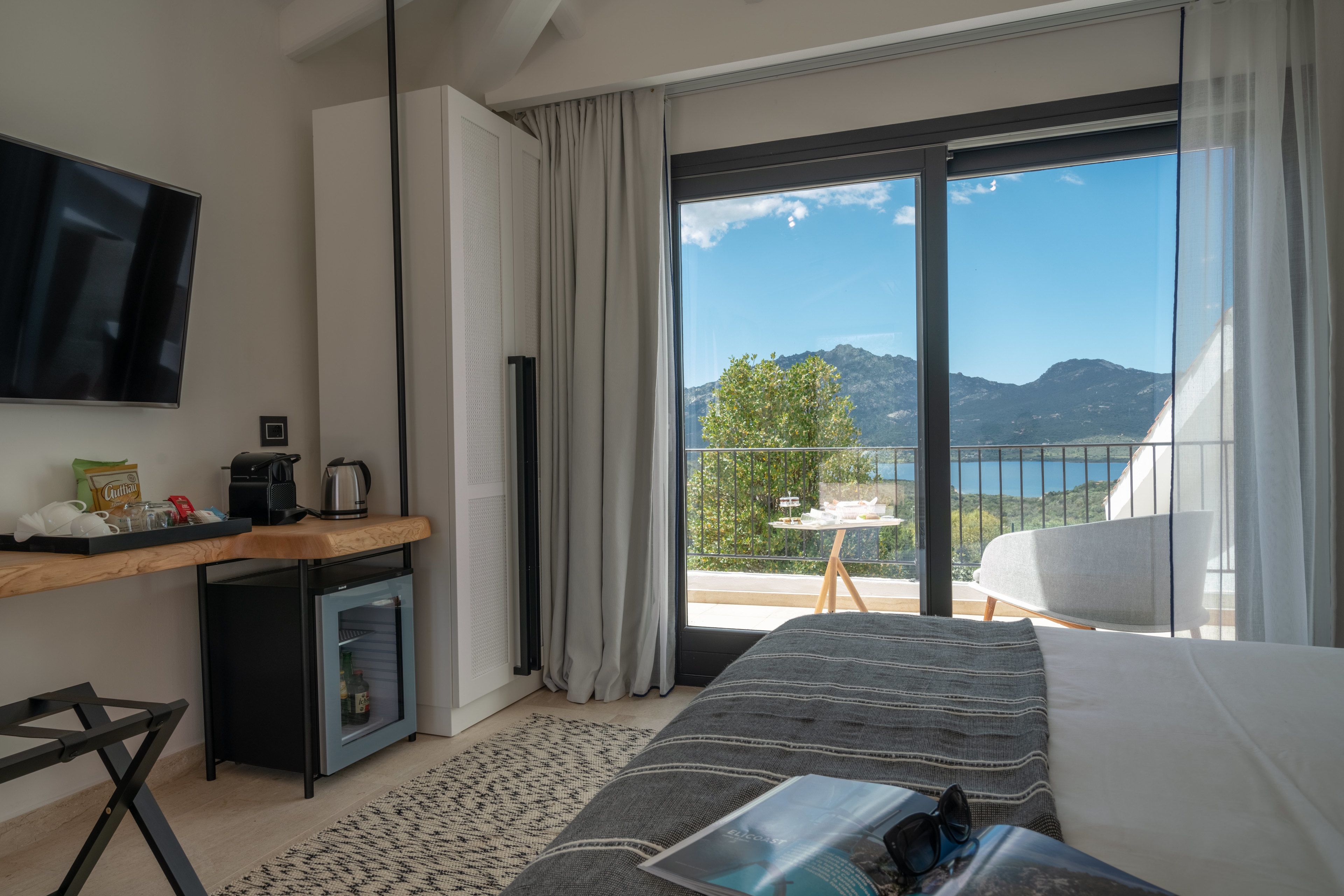A modern hotel room with a bed, a desk with a coffee maker, a mini-fridge, and a TV. The room has a balcony with a view of mountains and a lake.