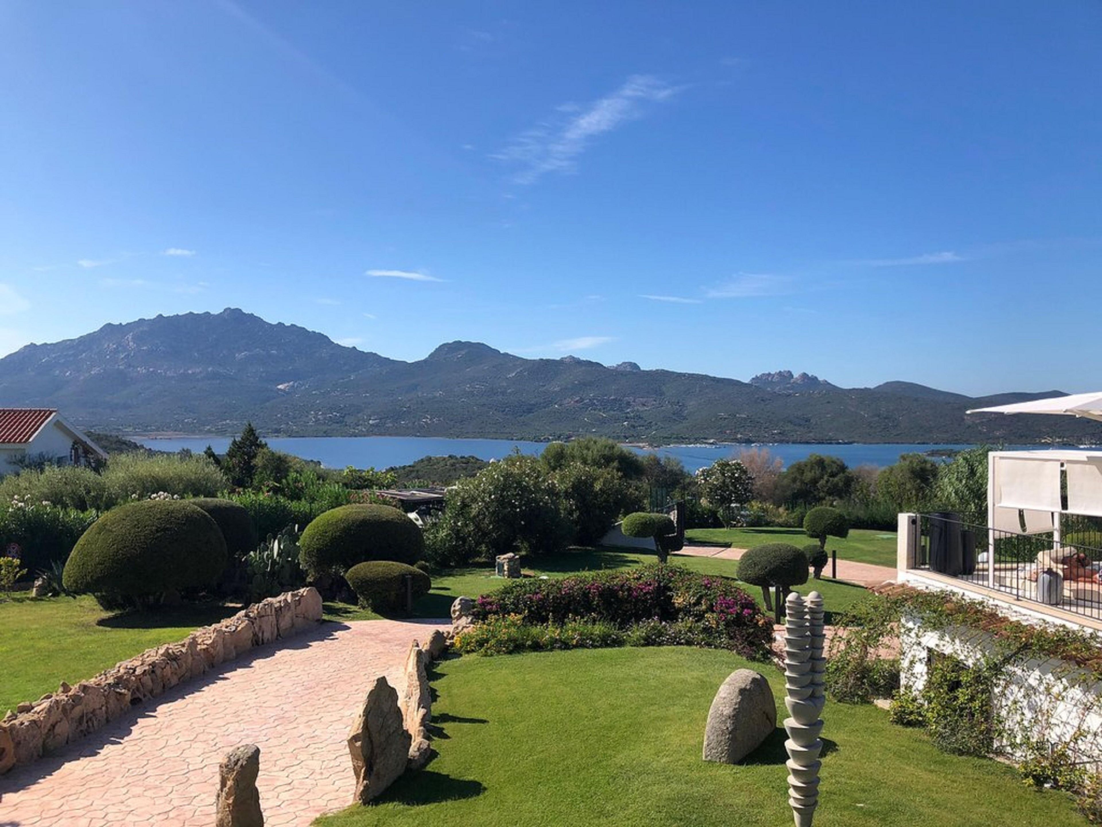A landscaped garden with a paved walkway, neatly trimmed bushes, and a view of a lake and mountains in the distance.