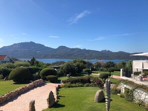 A landscaped garden with a paved walkway, neatly trimmed bushes, and a view of a lake and mountains in the distance. - Sulia House Porto Rotondo, Curio Collection by Hilton (Olbia)