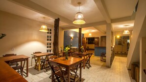 Un comedor con mesas y sillas de madera, iluminación colgante y vistas a la cocina al fondo.