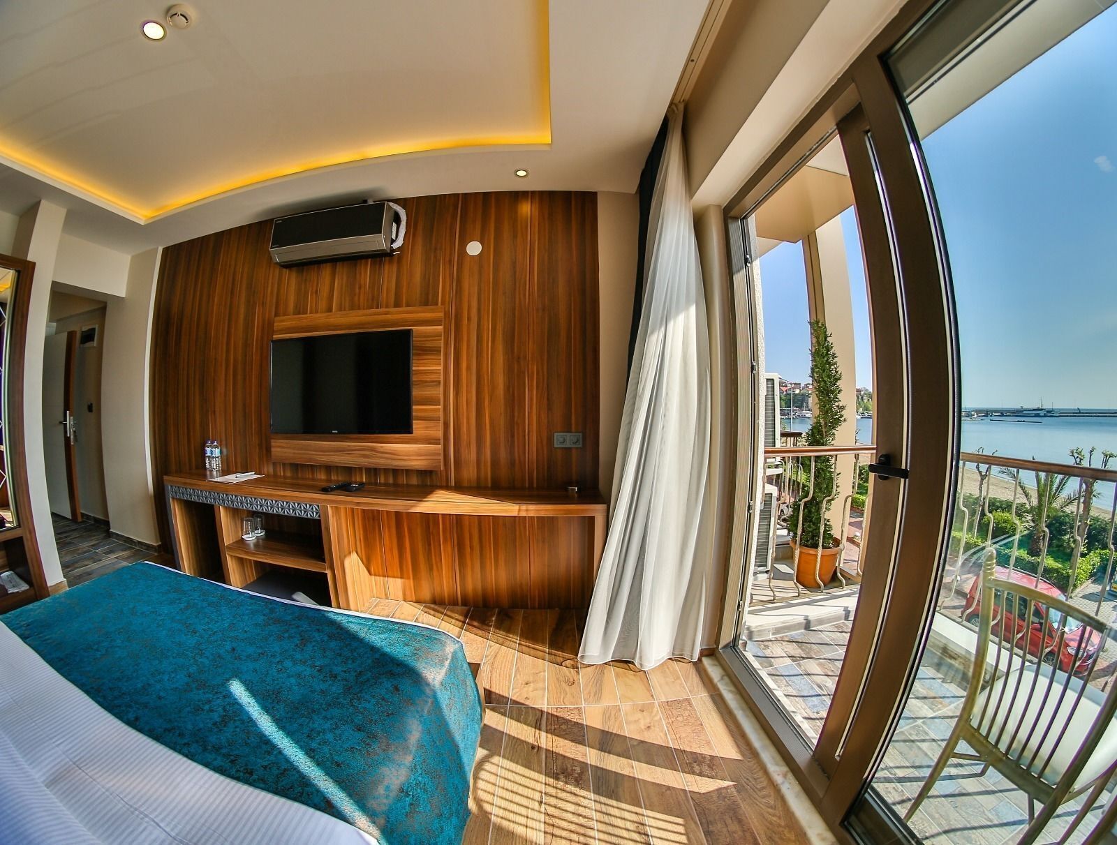 Une chambre d’hôtel moderne dotée d’une grande télévision, de murs recouverts de panneaux en bois, d’un balcon avec vue sur la mer et d’un lit avec une couverture bleue.