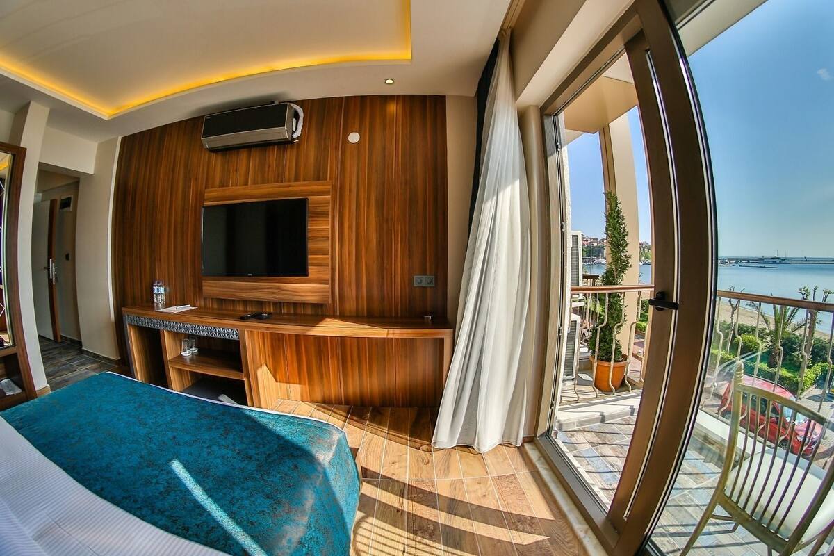 A modern hotel room with a large TV, wooden panel walls, a balcony with a view of the sea, and a bed with a blue cover.