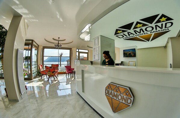 A reception area with a modern design, featuring a reception desk with a logo, a receptionist, and a view of the outdoors. - Diamond Liman Hotel (Amasra)
