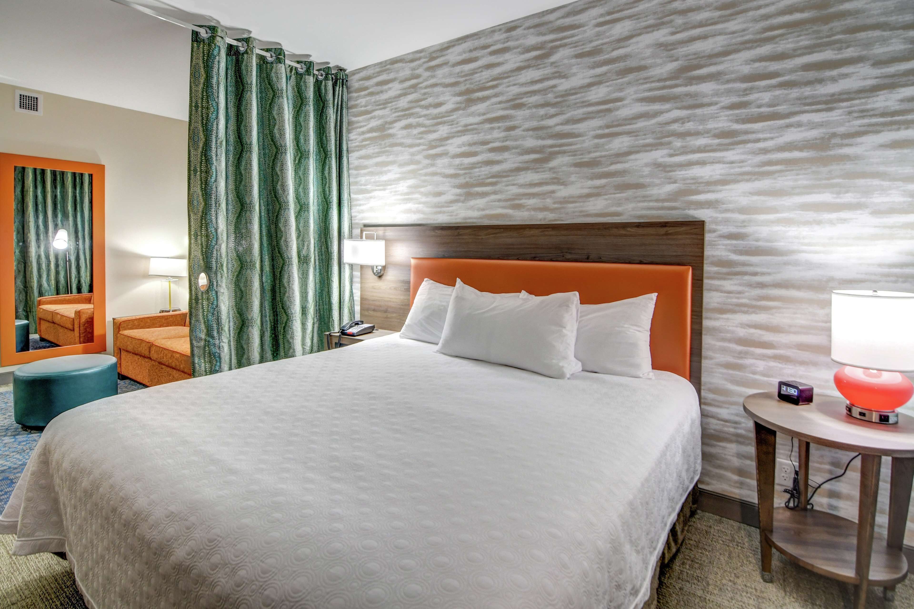 A hotel room with a large bed, a wooden headboard, a nightstand with a lamp, a chair, and a wall with a textured pattern.