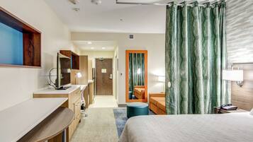 Une chambre d’hôtel comprenant un lit, un bureau, une chaise, un miroir, une salle de bain avec douche et donnant sur le couloir.
