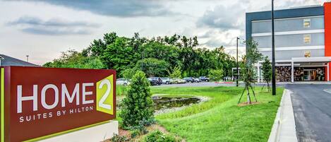 A hotel sign with "HOME 2 SUITES BY HILTON" and a landscaped area with grass and small trees.