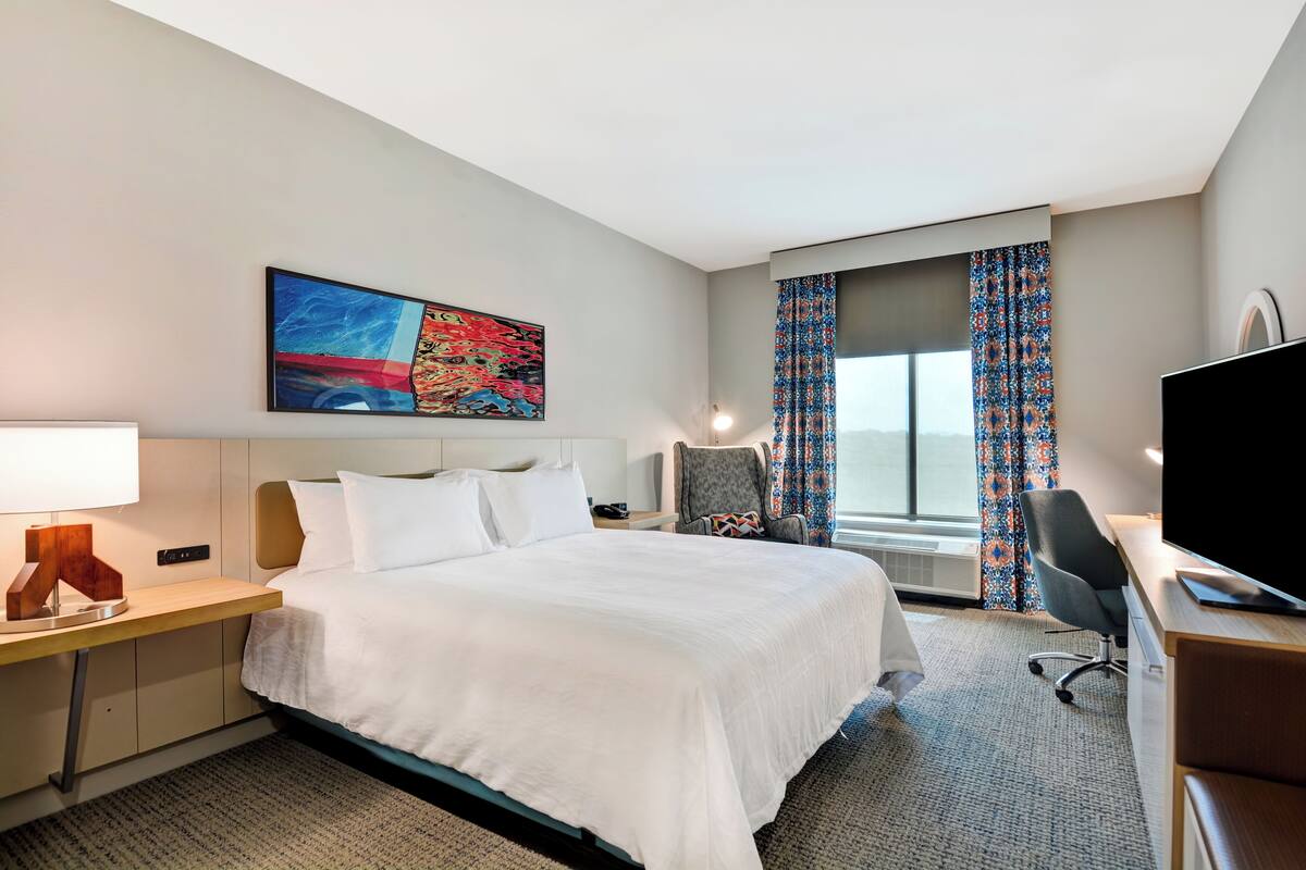 A hotel room with a bed, desk, chair, TV, and a painting on the wall.
