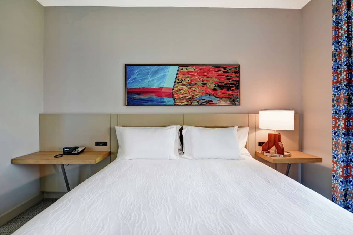 A hotel room with a large bed, two bedside tables, a phone, a lamp, and a colorful abstract painting on the wall.