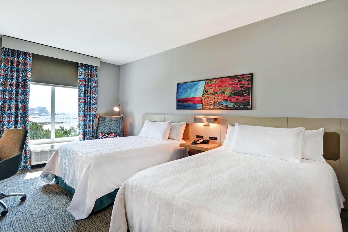 A hotel room with two beds, a chair, a desk with a phone, and a window with curtains.