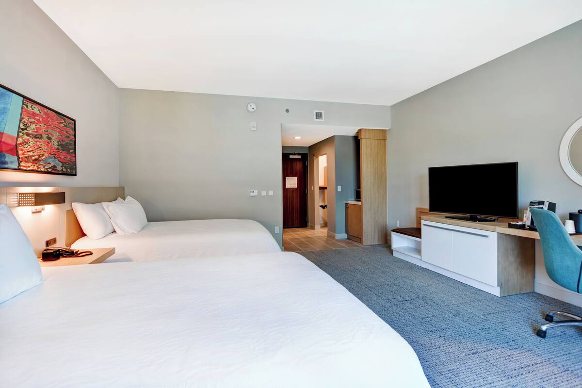 A hotel room with two beds, a TV, a desk with a chair, and a painting on the wall.