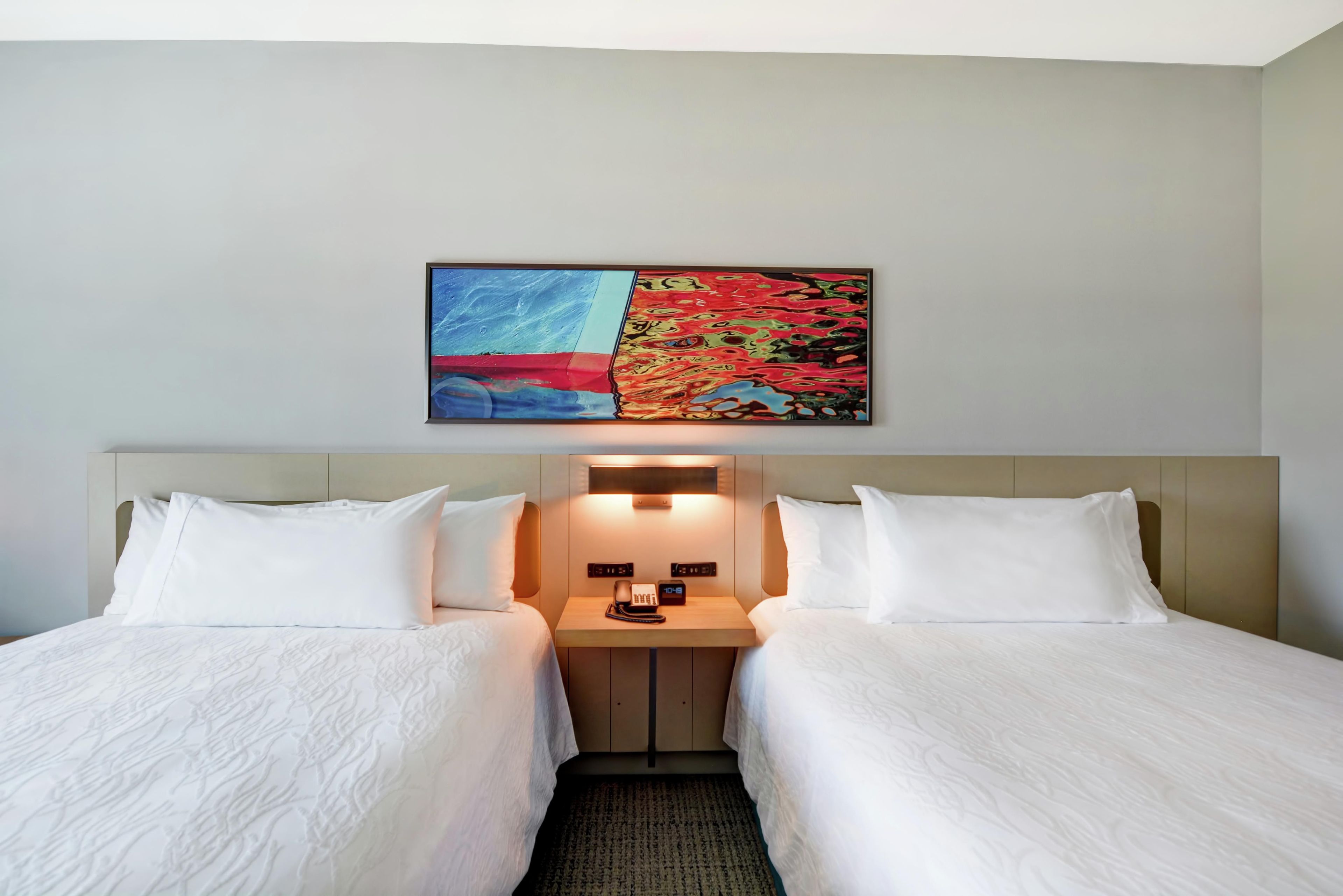 A hotel room with two beds, a nightstand with a phone, and a wall-mounted artwork.