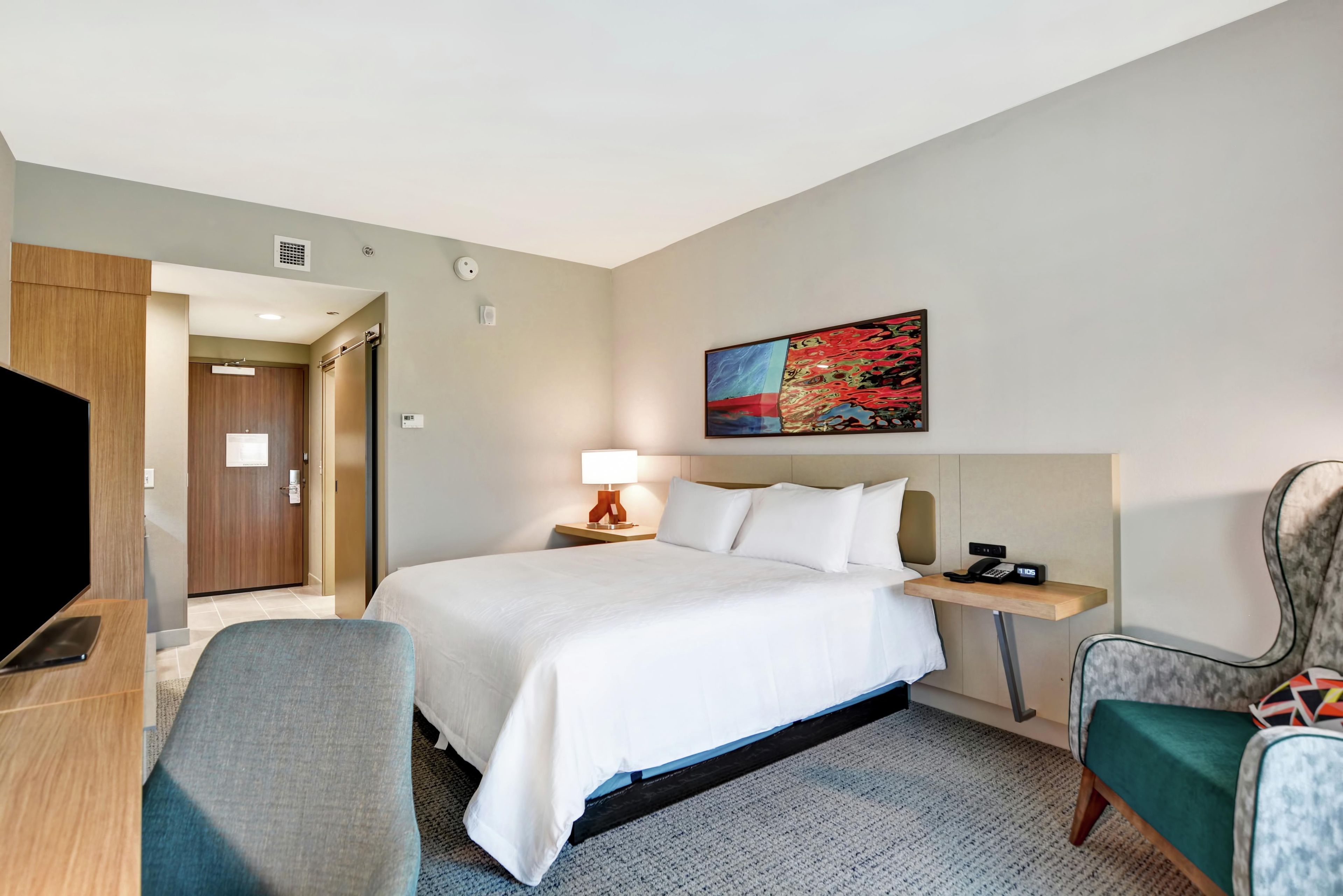 A hotel room with a bed, a desk, a chair, a nightstand, a lamp, a painting, and a telephone.