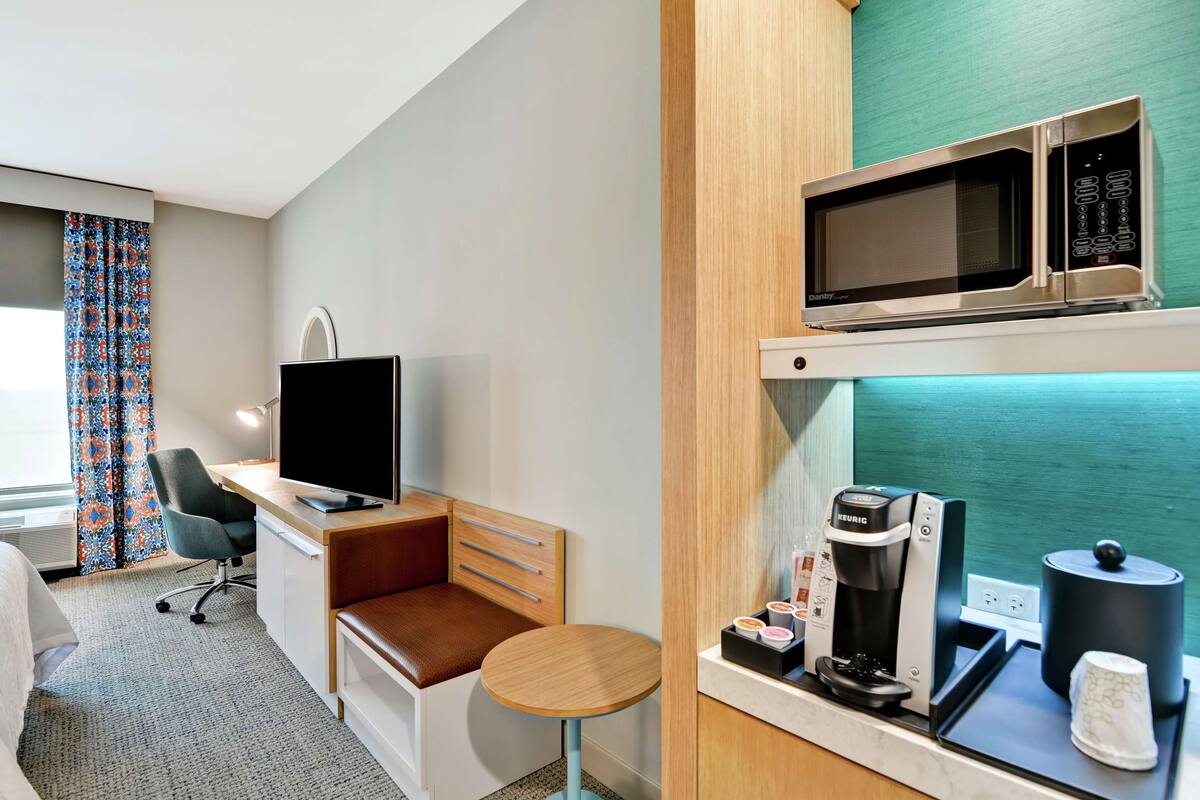 A compact hotel room with a bed, a desk with a computer, a TV, a coffee machine, and a microwave.