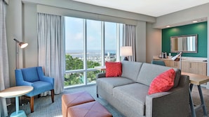 A modern hotel room with a large window, a grey sofa, a blue armchair, and a small round table. - Hilton Garden Inn Biloxi (Biloxi)
